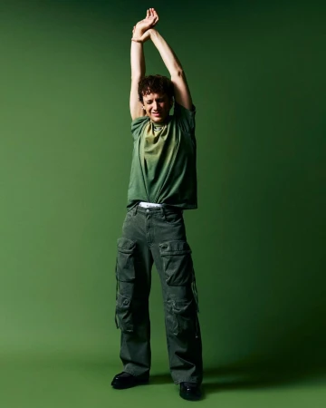 Full-body shot of a young adult man in green t-shirt and cargo pants, arms overhead, on a green background in a studio setting.