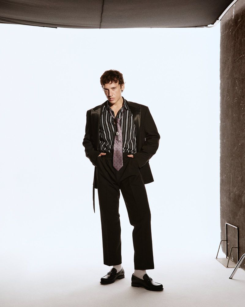A young adult man stands in a photo studio wearing a black suit and striped shirt with a tie, posing with his hands in his pockets.