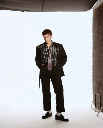 A young adult man stands in a photo studio wearing a black suit and striped shirt with a tie, posing with his hands in his pockets.