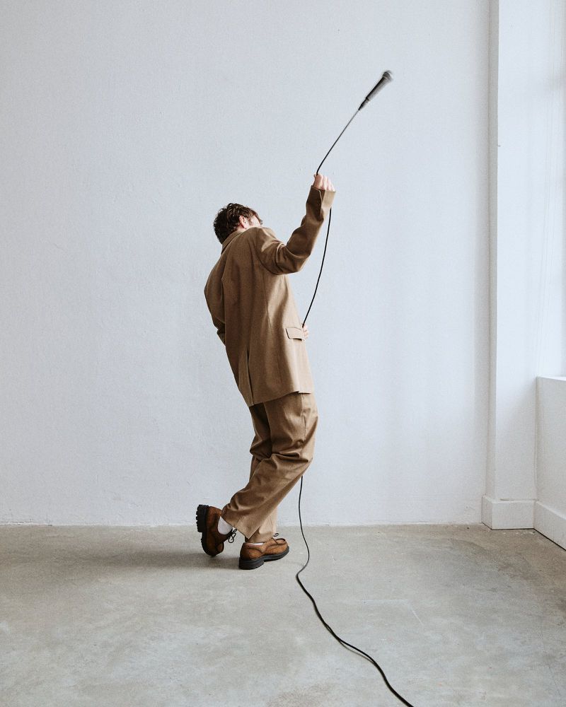 Adult in beige suit holds microphone cord in a white indoor space, wearing brown shoes, lit by natural light coming in from a window.