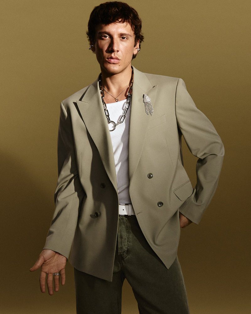 An adult male poses in a photo studio. He wears a sage green suit, white t-shirt, silver necklace, and olive pants against a neutral backdrop.
