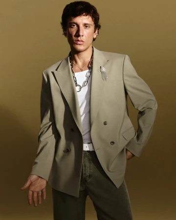 An adult male poses in a photo studio. He wears a sage green suit, white t-shirt, silver necklace, and olive pants against a neutral backdrop.