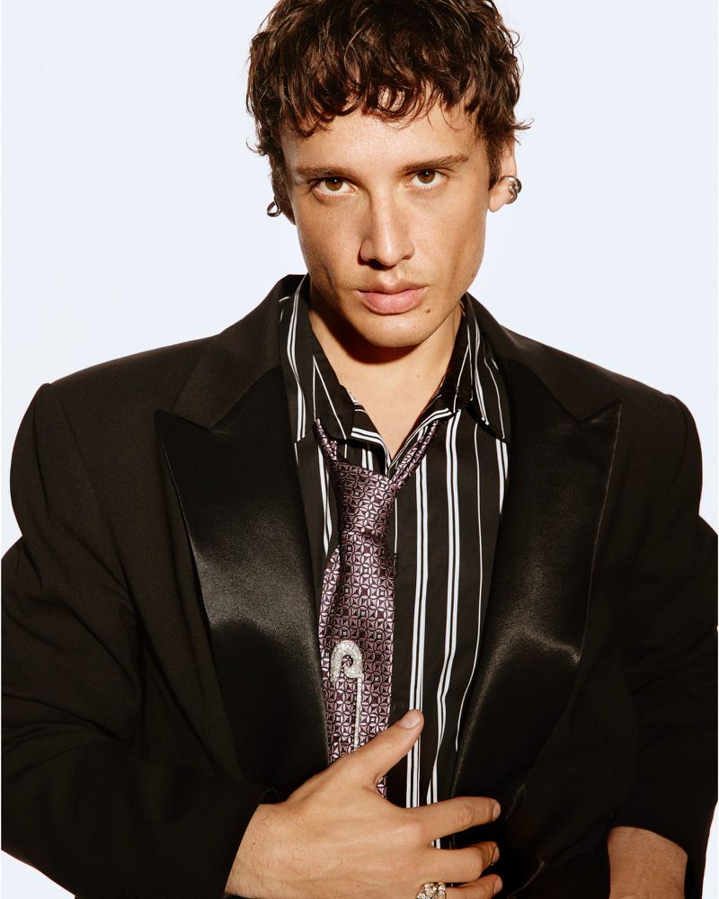Man in a black suit and striped shirt with a patterned tie, posing in a studio. Fashion-forward, stylish, and confident.
