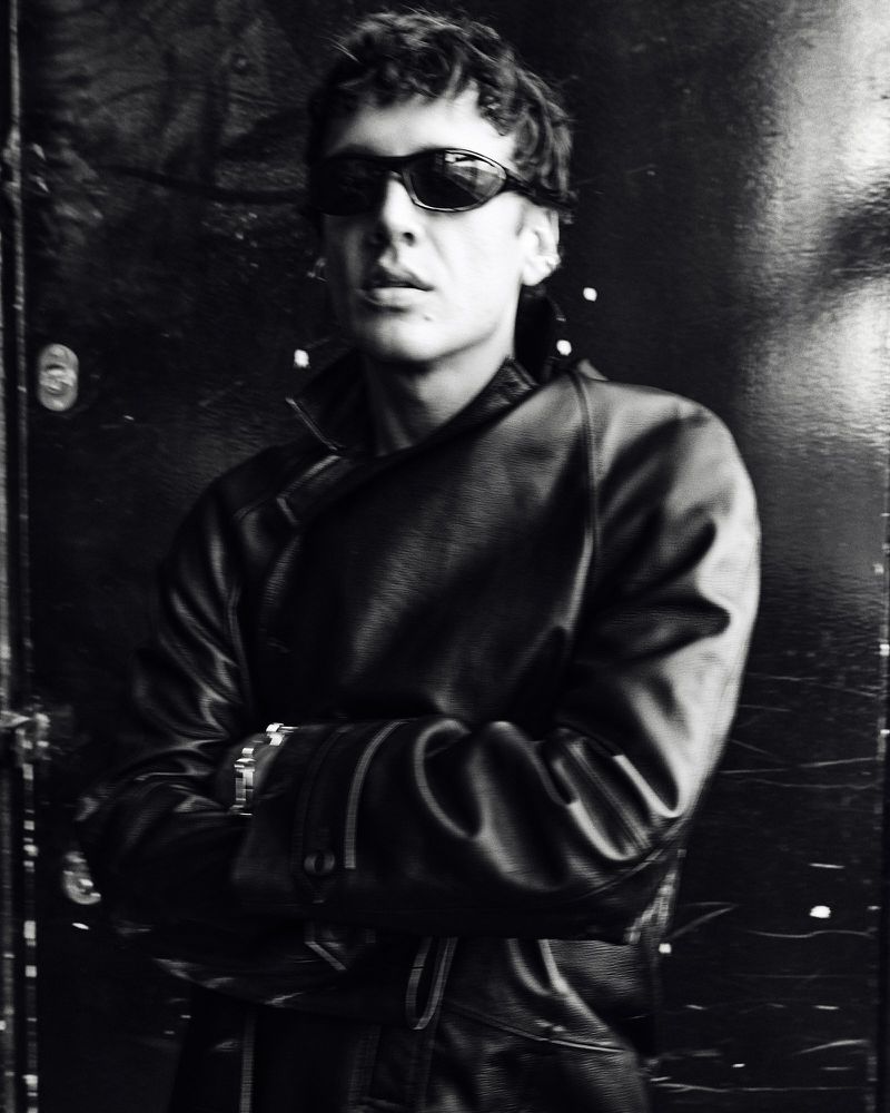 Man wearing sunglasses and a black leather jacket with arms crossed, photographed in monochrome.