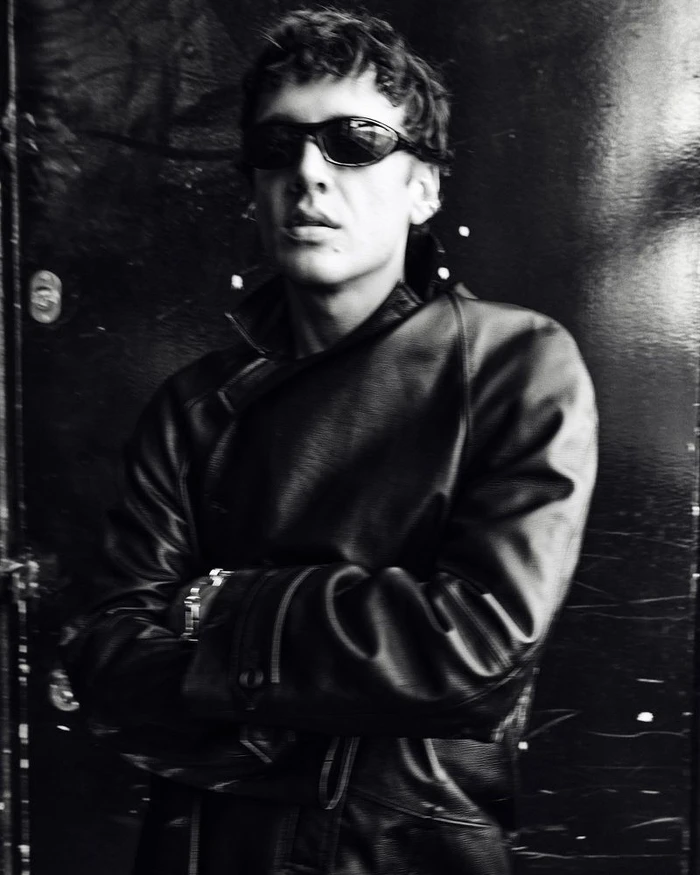 Man wearing sunglasses and a black leather jacket with arms crossed, photographed in monochrome.