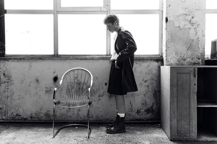 Monochrome fashion editorial image of a young man standing next to a metal wire chair in an industrial room with large windows.