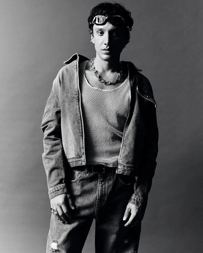 Man wearing a denim jacket and mesh tank top with sunglasses pushed onto his forehead against a grey background