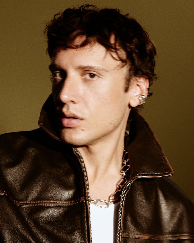 Portrait of a man wearing a dark leather jacket and silver chain necklace against an olive green background