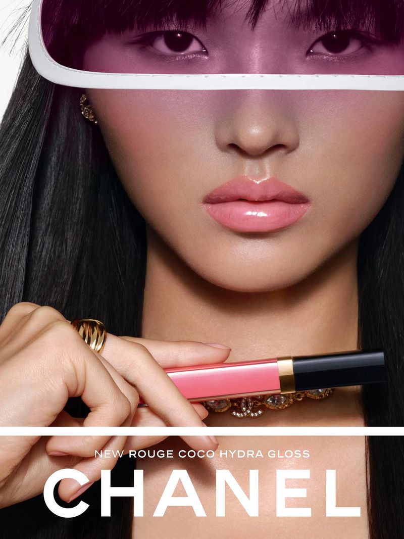 Model Wei Yiting wearing a translucent pink visor while holding a pink Chanel lip gloss tube horizontally against a white studio backdrop.
