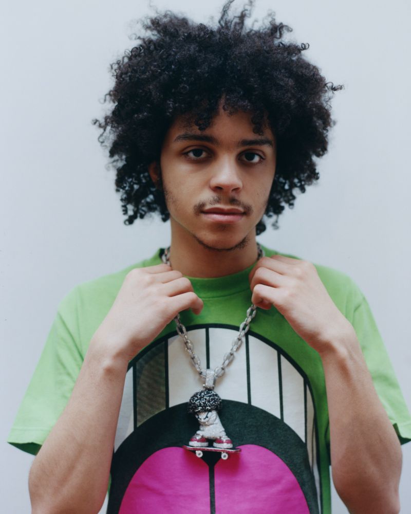 Portrait of musical artist xaviersobased wearing a green graphic t-shirt and custom skateboard pendant against a white background.