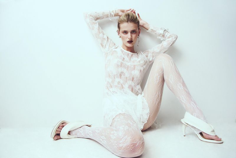 A woman in a white lace outfit and heels poses in front of a solid white backdrop for a fashion editorial photoshoot.