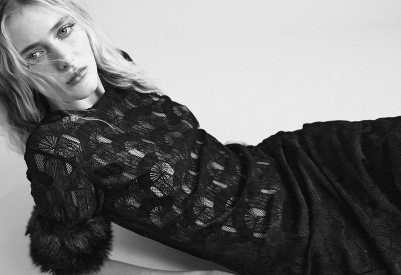 A fair-skinned woman is posed against a white background, wearing a long-sleeved black lace dress with fur cuffs in a studio setting.