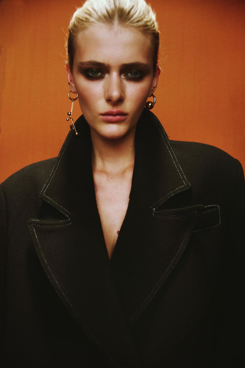 Close-up fashion shot of a fair-skinned woman with blonde hair and heavy eye makeup wearing an oversized black jacket indoors.