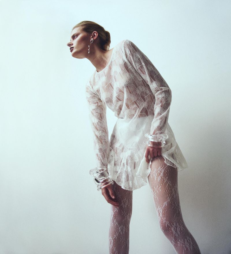 A striking fashion shot of a solitary woman wearing a white lace top, mini skirt, and fishnet stockings against a white backdrop.