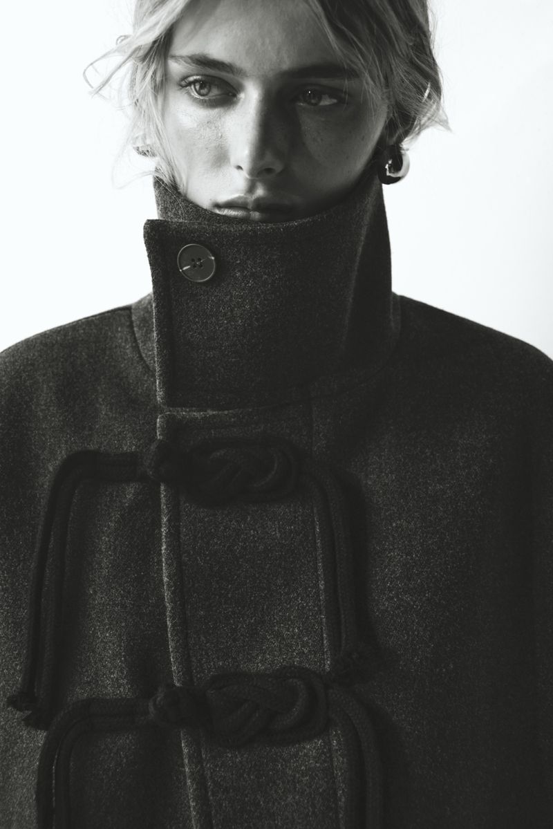 Monochrome portrait of a woman in a high-collared grey coat with rope details and button, isolated on a white background.