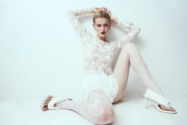 Model Cara Fravel seated wearing a sheer white lace dress and white heels against a clean white backdrop for Tidal Magazine.
