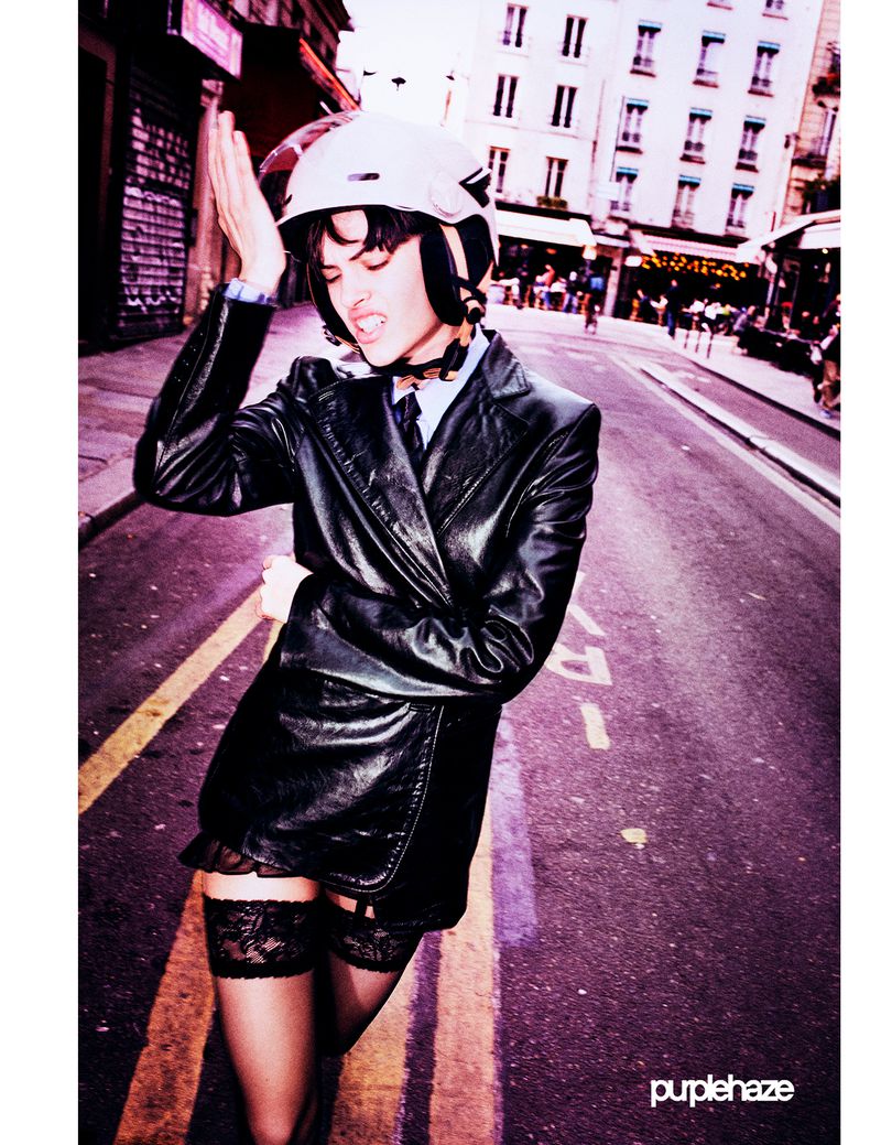 Adult female on urban street wears a black leather jacket, mini dress with lace trim, and a white helmet; city buildings in the background.
