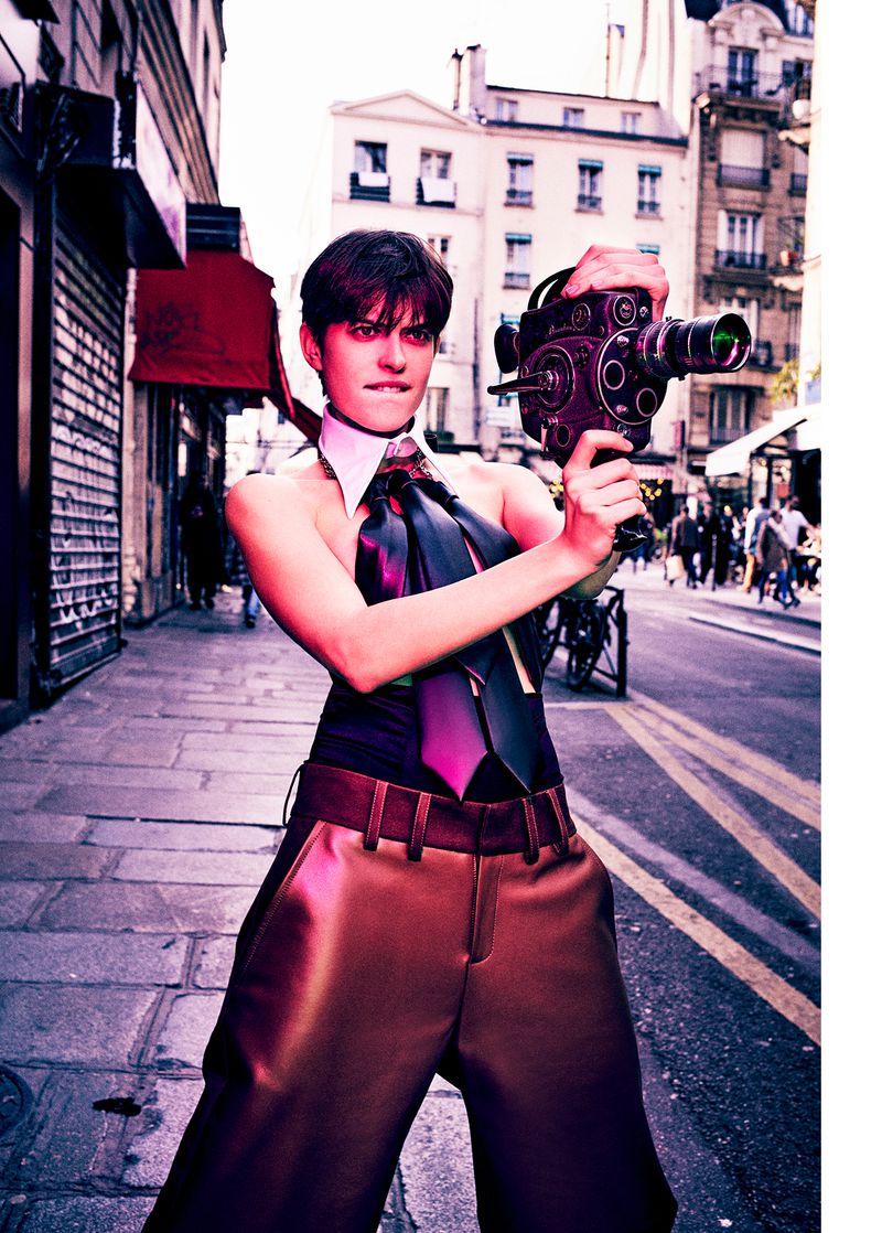 Fashion editorial shot of a woman holding an antique film camera on a European city street, posing in oversized brown pants and a collared top.