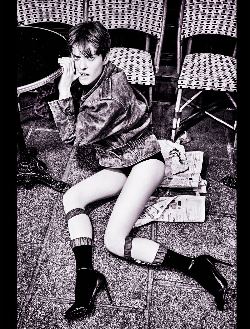Fashion photo of a woman in a jacket, shorts, and high heels lying on a patterned stone floor near chairs and a table.
