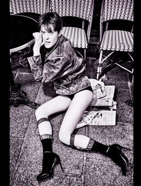 Fashion photo of a woman in a jacket, shorts, and high heels lying on a patterned stone floor near chairs and a table.