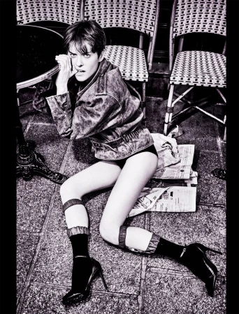 Fashion photo of a woman in a jacket, shorts, and high heels lying on a patterned stone floor near chairs and a table.