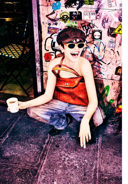 Woman in sunglasses and cap laughs while squatting outdoors with a beverage and cigarette near a graffiti-covered wall and outdoor cafe furniture.