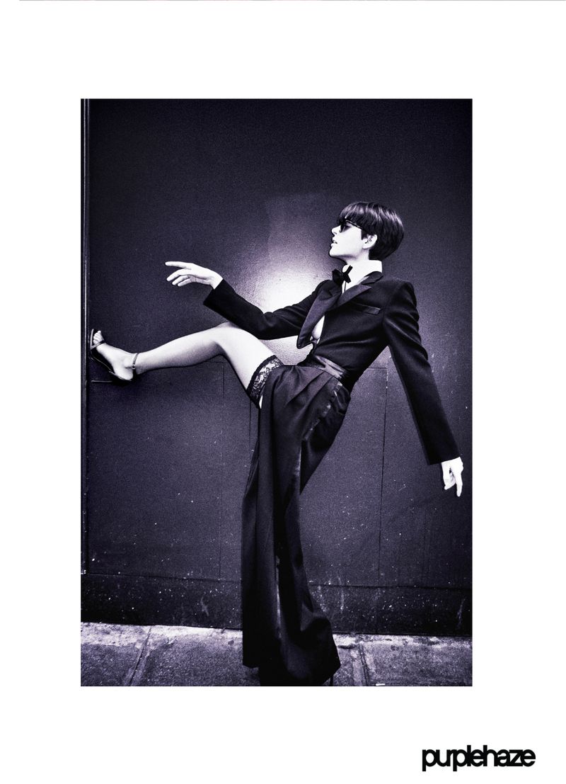 Fashionable woman in a black tuxedo and lace garter strikes a pose against a dark wall in an artistic studio shot, vintage-inspired.