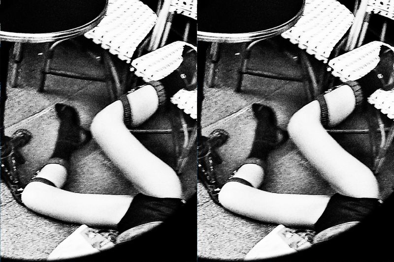 Black and white shot of legs and feet, in socks and shoes, crossed under a table with a patterned bistro chair in the background.