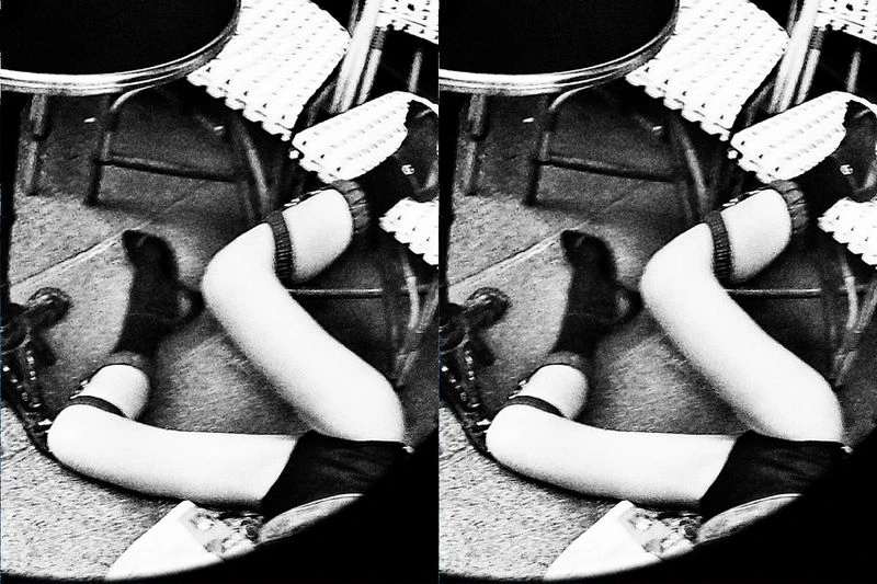 Black and white shot of legs and feet, in socks and shoes, crossed under a table with a patterned bistro chair in the background.