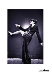 Model in a black tuxedo jacket and high-waisted trousers lifting one leg against a dark wall in a monochrome editorial photograph