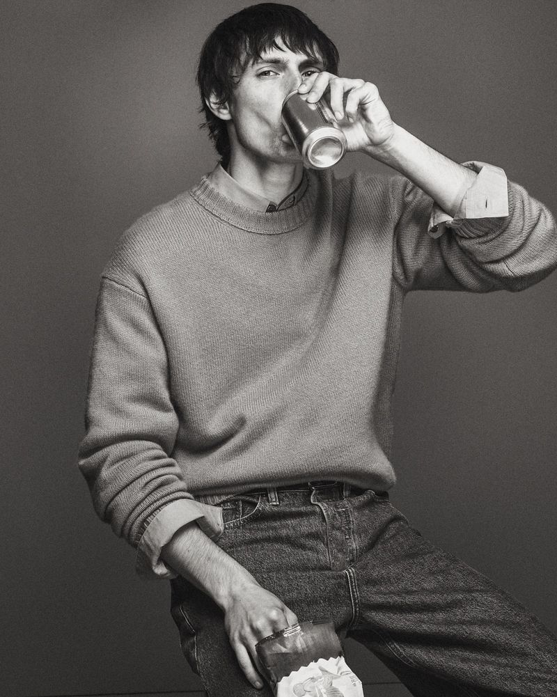 Model drinking from a can while wearing a knit sweater and denim jeans against a solid dark background