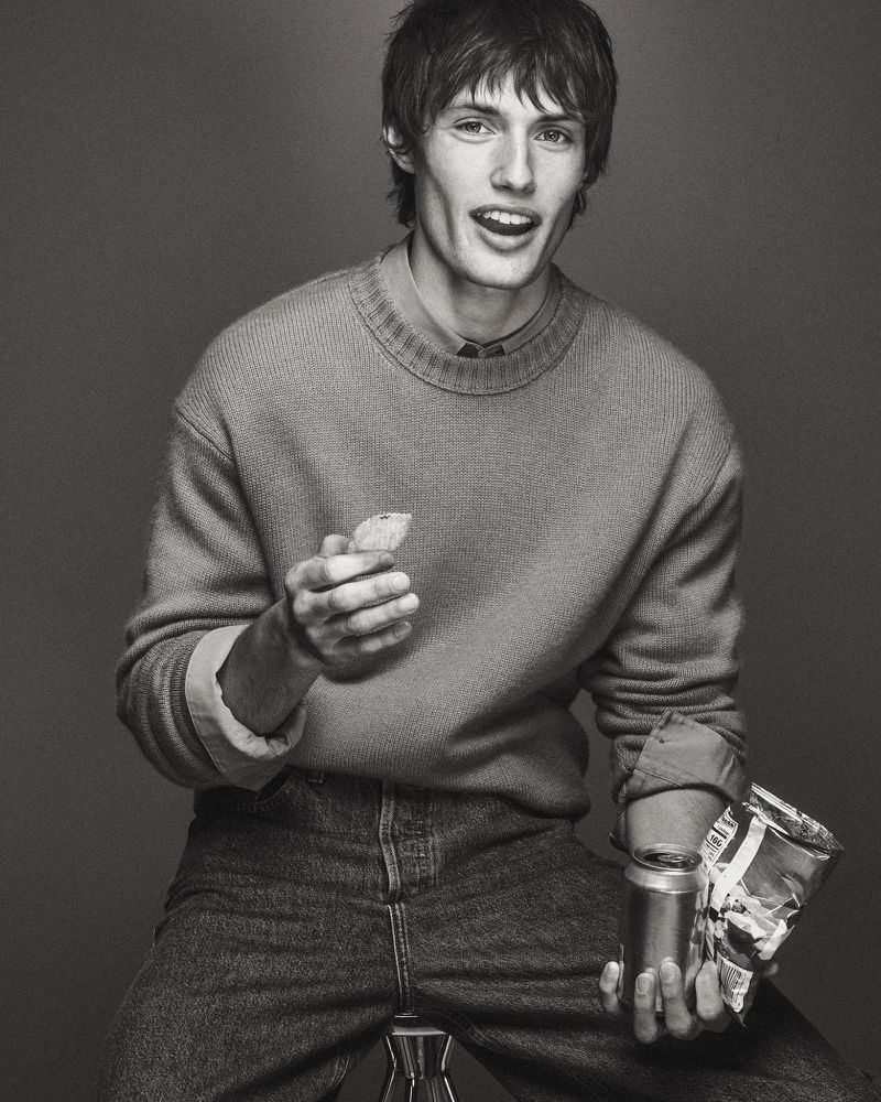Monochrome studio portrait of model Thatcher Thornton holding a chip and beverage can wearing a knit sweater and jeans.