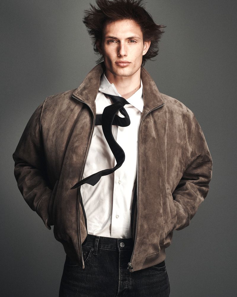 Thatcher Thornton wearing a brown suede bomber jacket, white shirt, floating black tie, and dark jeans against a grey studio backdrop.