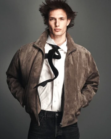 Thatcher Thornton wearing a brown suede bomber jacket, white shirt, floating black tie, and dark jeans against a grey studio backdrop.