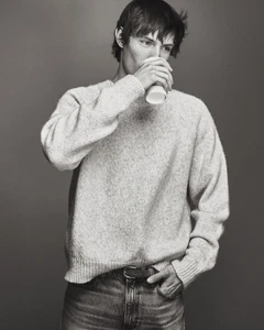 Model Thatcher Thornton drinking from a paper cup while wearing a knit sweater and jeans against a neutral studio backdrop.