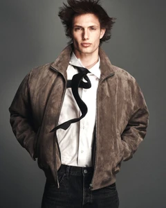 Thatcher Thornton wearing a brown suede bomber jacket, white shirt, floating black tie, and dark jeans against a grey studio backdrop.