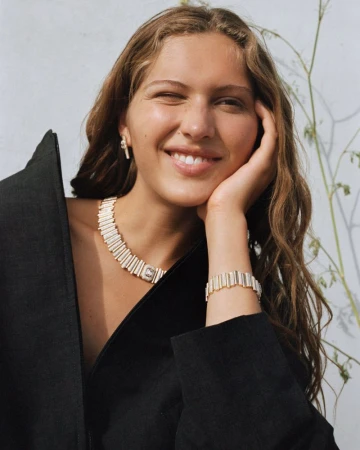 Model Angelina Kendall winks and smiles while wearing a structured black jacket and ribbed gold statement jewelry.