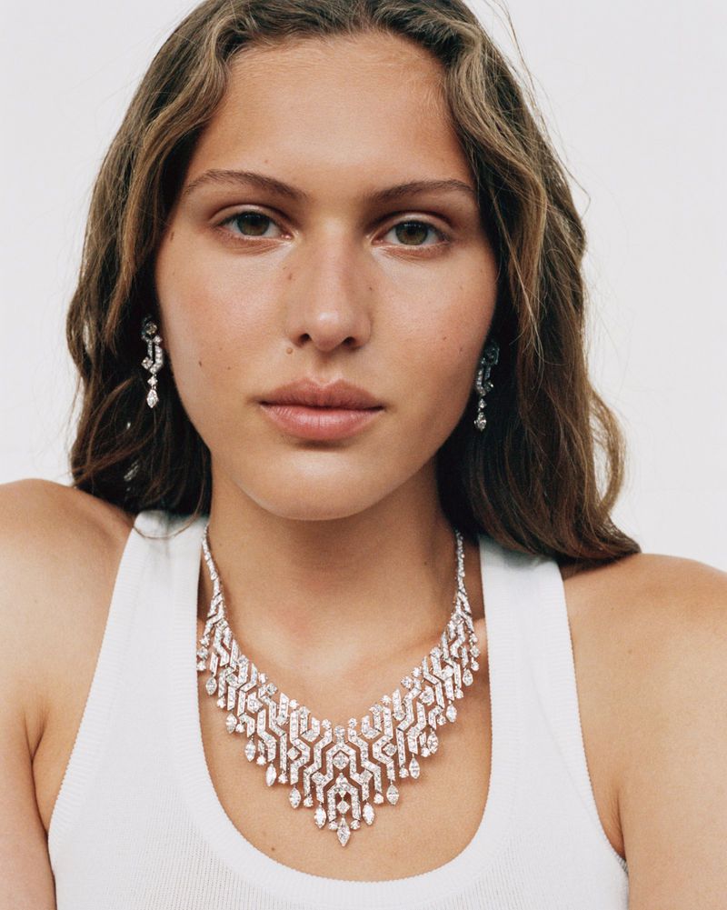 Woman with light brown wavy hair wearing white tank top, diamond earrings, and a geometric diamond necklace against a white backdrop.