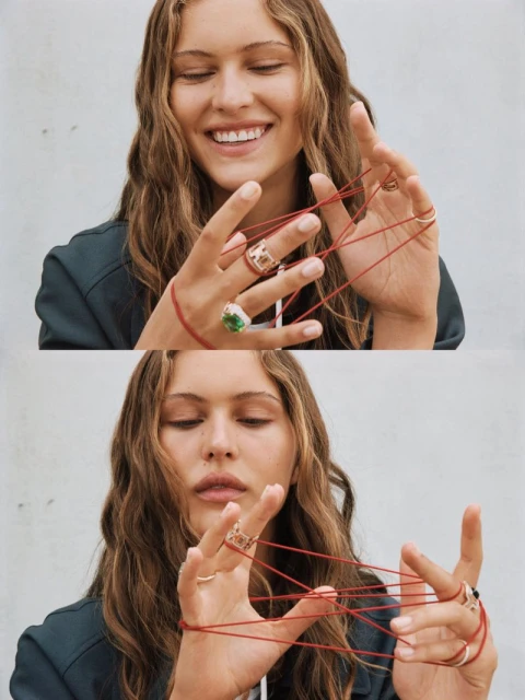 Smiling woman plays cat's cradle with red string on her fingers. She wears rings and a dark jacket against a neutral wall backdrop.