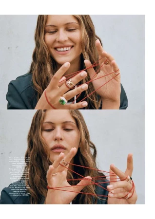 Close-up shot of a smiling young woman with long wavy hair, wearing multiple rings, playing with a red string indoors.