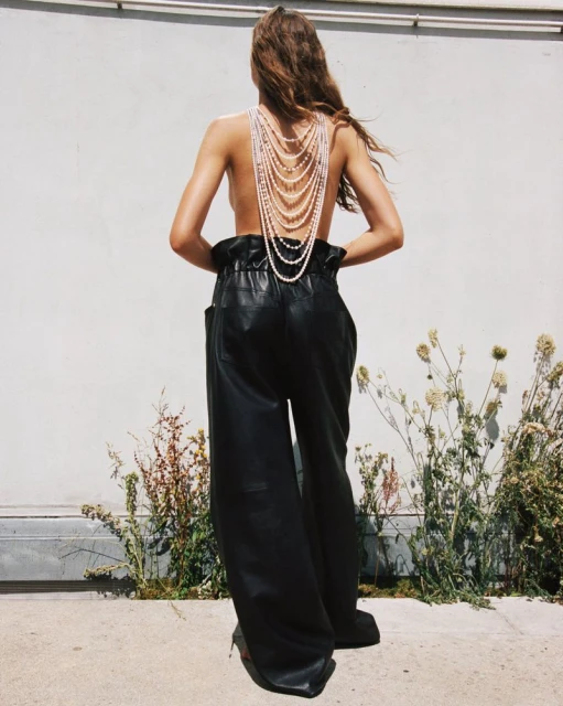 Back view of a slender woman with long hair wearing black leather pants and multiple strands of pearls standing outdoors.
