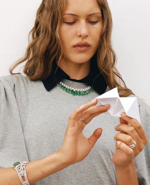 Woman in grey t-shirt wears diamond and emerald jewelry and holds a paper fortune teller with a white studio background.