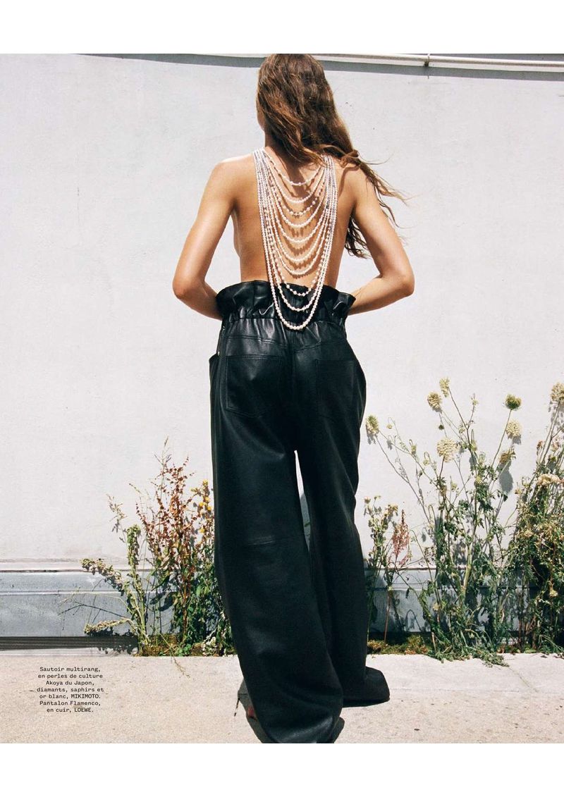 Back view of a woman wearing black leather pants and a pearl necklace standing outside with bright sunlight casting shadows.