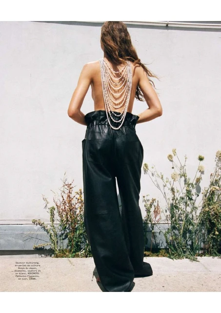 Back view of a woman wearing black leather pants and a pearl necklace standing outside with bright sunlight casting shadows.
