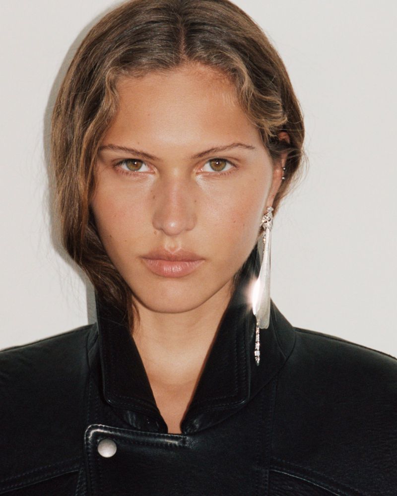 Close-up studio portrait of a woman with light brown hair and a black leather jacket, wearing a long silver statement earring.