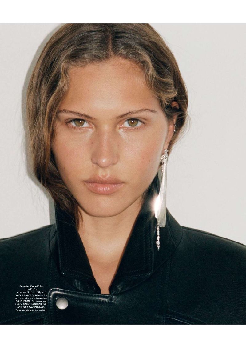 Close-up shot of a young adult woman with wavy brown hair wearing a black leather jacket and long silver earrings against a white backdrop.