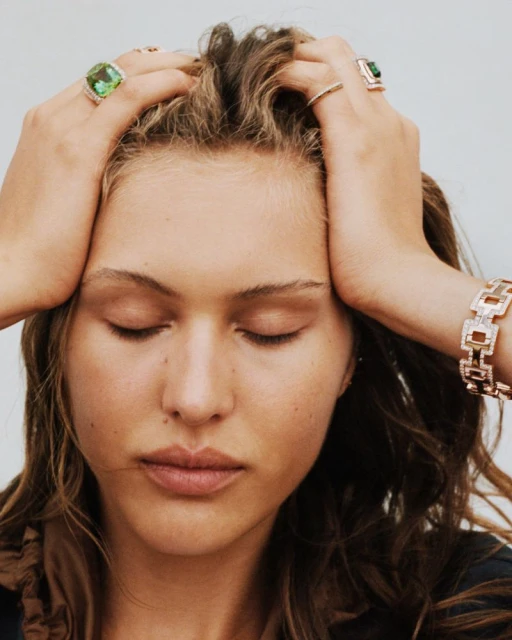 A woman with closed eyes touches her head, showcasing statement jewelry rings and bracelet in a close-up studio shot against a white backdrop.