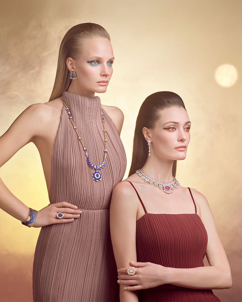 Two women posing in pleated dresses wearing high jewelry necklaces, rings, and earrings against a golden gradient background.