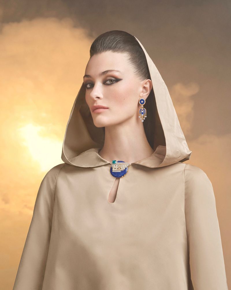 Model wearing a beige hooded cape and ornate blue and gold lapis lazuli jewelry against a warm sunset sky.