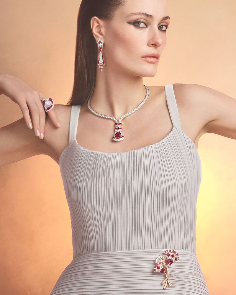 Model Jace Higgins posing in a pleated grey dress adorned with diamond and ruby high jewelry against a warm studio backdrop.